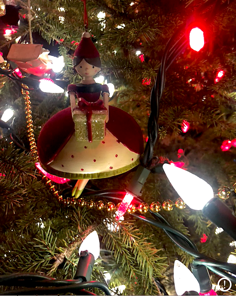 A christmas ornament of a woman in an old style dress holding a gift, tree branches and lights surround her.