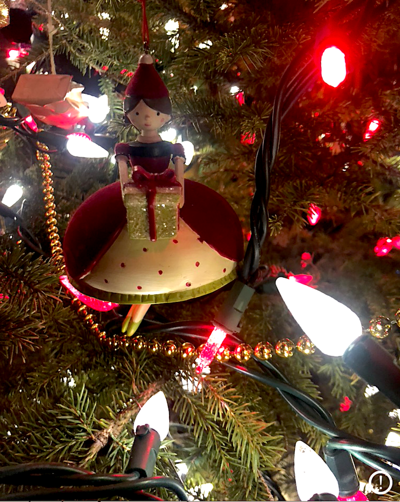 A christmas ornament of a woman in an old style dress holding a gift, tree branches and lights surround her. 