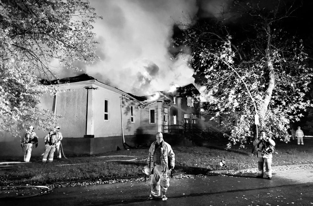 Men in fire fighting gear in front of burning building. 