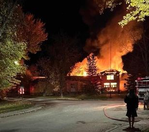 A person in shadows stands in front of a burning building (a school). 