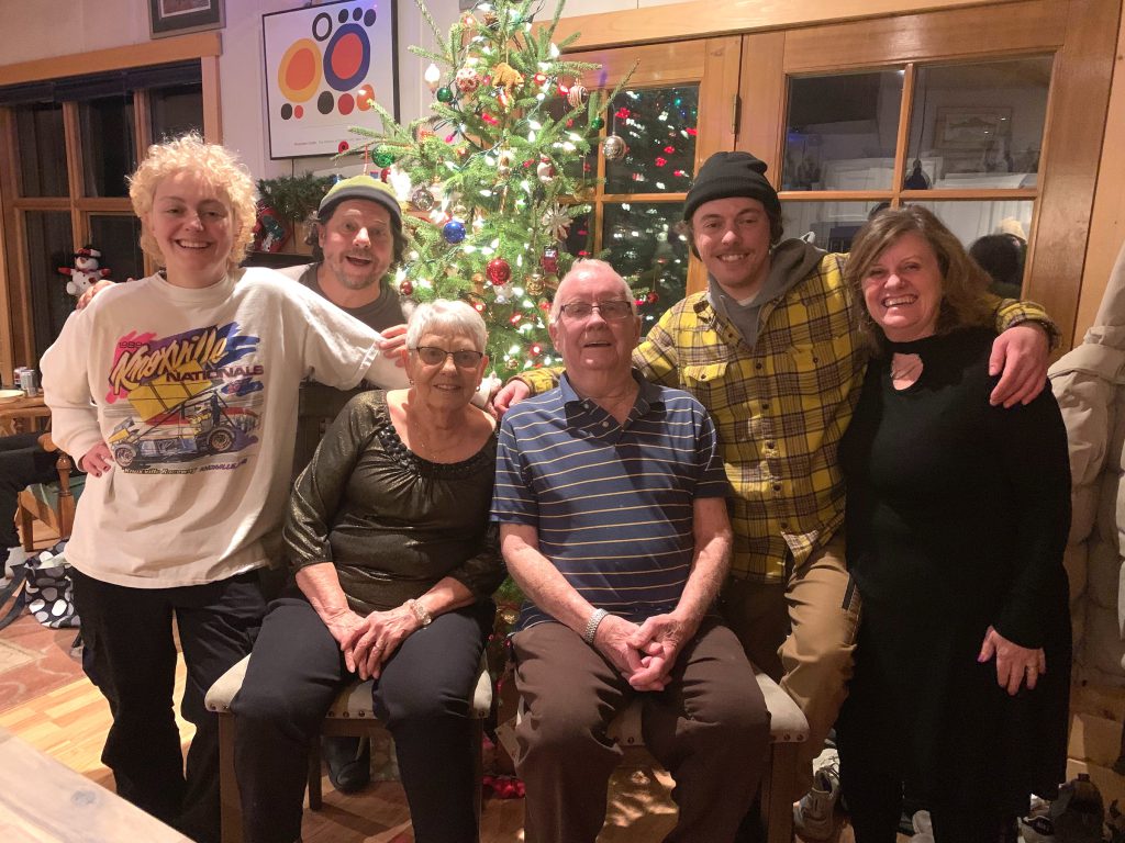 Family of six sitting in front of a Christmas tree, two older grandparents,  parents and adult children. 