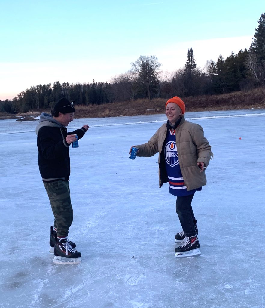 two young people on an outdoor skating rink,in skates, winter clothes, one in an Edmonton oiler jersey, both holding beverages.