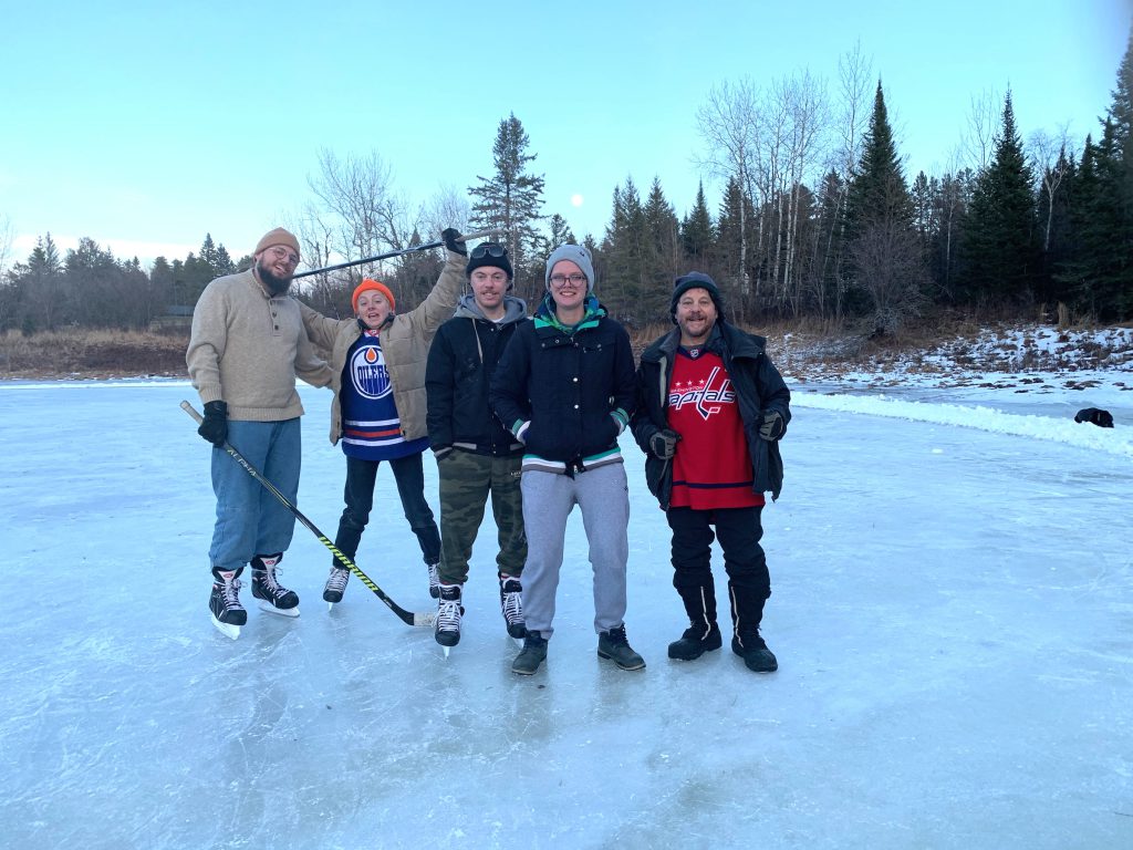 five people on an outdoor rink, one is holding a hockey stick above their head, 3 are in skates and 2 in boots. 