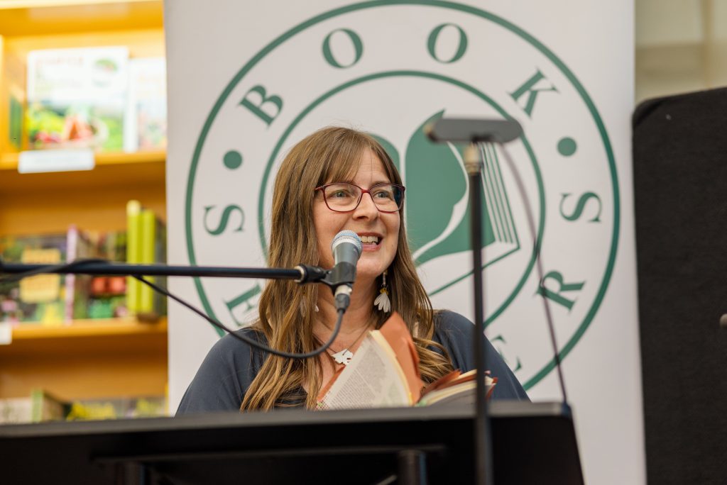 Angeline Schellenberg with long brown hair, wearing red glasses is holding a book and speaking into a microphone.
