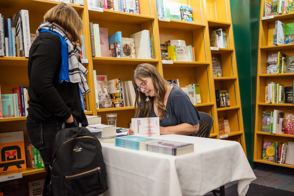 Angeline Schellenberg is in a blue dress and sitting at a table and signing books. A book buyer dressed in black and holding a black backpack is standing in front of the table.